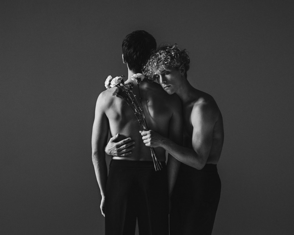 A black and white photo of a blonde man holding carnations while embracing a dark-haired man whose back is to the camera.